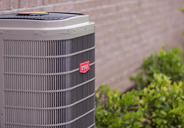 Bryant brand heat pump located on the side of a brick house, next to bushes.
